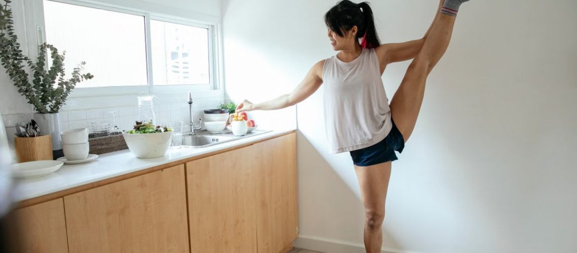 Full length of young female in sleepwear standing in kitchen and doing splits with leg up near counter while making tea for meal