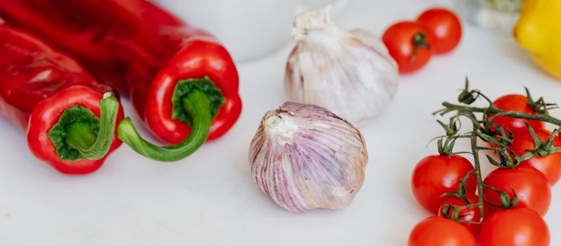 Fresh garlic placed among ripe red cherry tomatoes and peppers on white table near white ceramic cup and yellow lemon