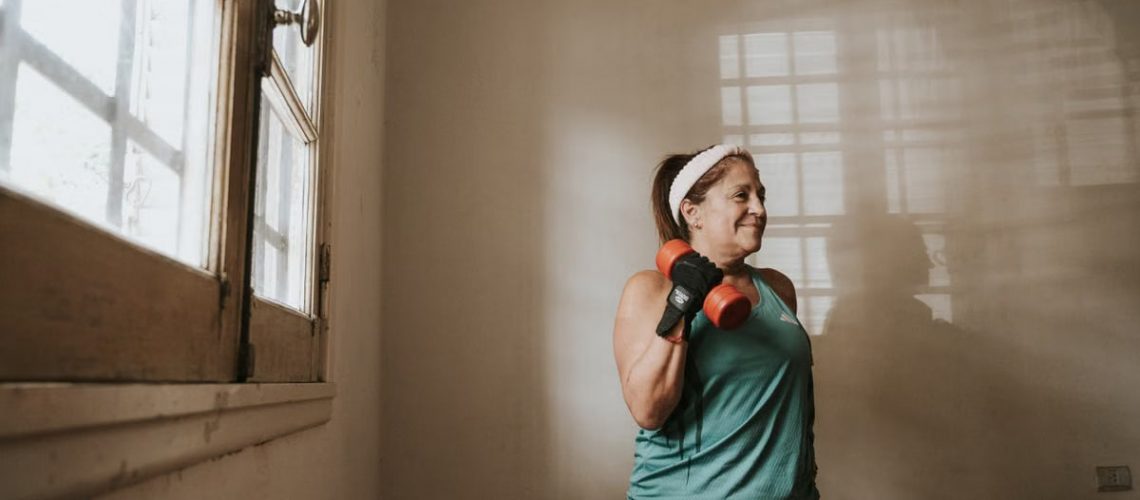 Senior woman in a brightly lit room exercises with dumbbells, promoting fitness at home.